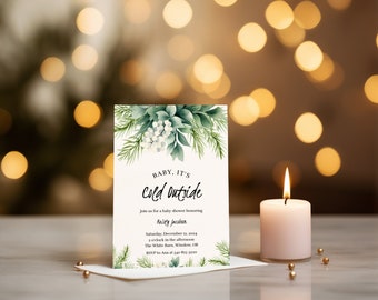 May include: A baby shower invitation with the text "BABY, IT'S cold outside" and floral accents. The invitation includes details for a baby shower honoring Kristy Jacobson, with a candle and gold beads in the foreground.