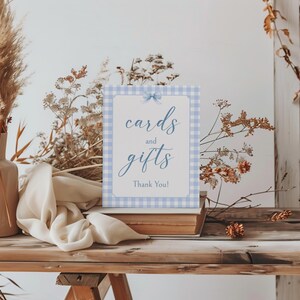 May include: A blue and white gingham sign with the text "cards and gifts" and "Thank You!" in blue script. The sign is propped up on a stack of books on a wooden table.