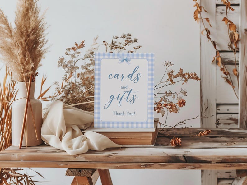May include: A blue and white gingham sign with the text "cards and gifts" and "Thank You!" in blue script. The sign is propped up on a stack of books on a wooden table.
