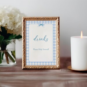 May include: A gold framed sign with a blue and white gingham pattern and a blue bow. The sign says "drinks" and "Please Help Yourself".