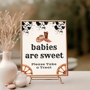 May include: A white sign with black text that reads "babies are sweet" and "Please Take a Treat". The sign is decorated with a watercolor illustration of cowboy boots and a cowboy hat. The sign is on a gold stand.