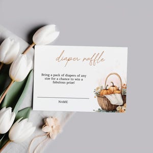 May include: A white card with the words "diaper raffle" in gold script. The card includes text inviting guests to bring diapers for a prize, and a watercolor illustration of pumpkins in a basket. White tulips and greenery are in the background.