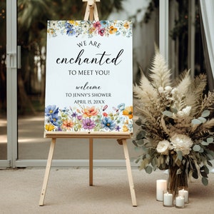May include: A white welcome sign with floral border and the text "We are enchanted to meet you! Welcome to Jenny's Shower April 15, 20XX". The sign is on an easel in front of a bouquet of dried flowers.