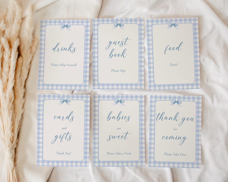 May include: Six blue and white gingham patterned signs with blue ribbon accents and white text. The signs read "drinks", "guest book", "food", "cards and gifts", "babies are sweet", and "thank you for coming".