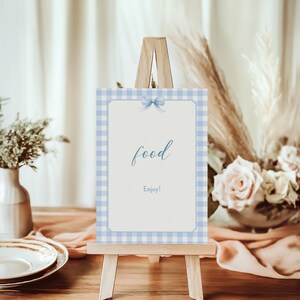 May include: A blue and white gingham sign with a blue bow on a wooden easel. The sign says "food" and "Enjoy!"