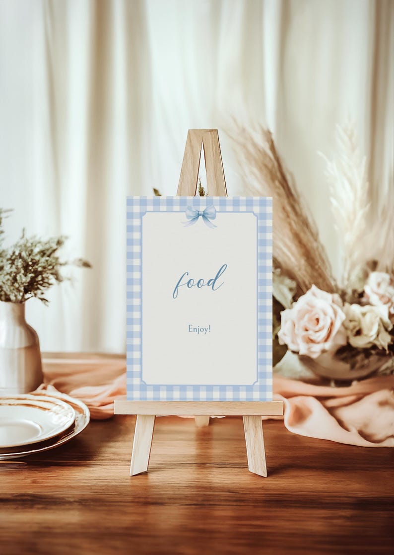 May include: A blue and white gingham sign with a blue bow on a wooden easel. The sign says "food" and "Enjoy!"