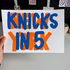 Op de afbeelding: Een rechthoekig wit papieren bord met de handgeschilderde oranje tekst NEW YORK met de blauwe tekst KNICKS IN 5 eroverheen. De belettering is vet en blokvormig, gebruikmakend van een levendig kleurenschema van feloranje en diep koningsblauw.