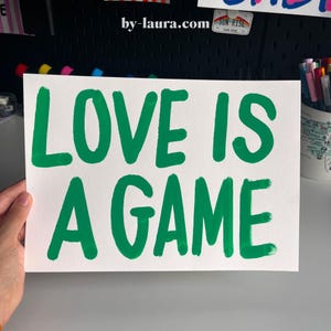 May include: A white paper with the phrase "LOVE IS A GAME" in bold, green letters. The artwork is held by a hand, with the website by-laura.com visible in the background. The overall theme is a motivational or artistic statement.