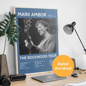 Puede incluir: Un póster azul y blanco con una foto de un hombre tocando la guitarra. El póster se titula "Mark Ambor: The Rockwood Tour" y enumera las fechas y lugares de la gira. El póster está sobre una mesa de madera con una computadora portátil, una lámpara y una planta.