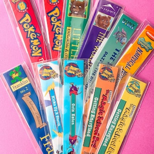 Puede incluir: Una colección de coloridos marcadores, cada uno en una funda de plástico transparente. Presentan personajes de dibujos animados y títulos como "Pokémon" y "The Magic School Bus", con diseños vibrantes y texto.
