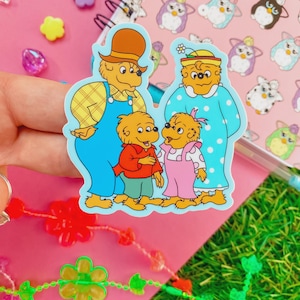 May include: A colorful sticker featuring the Berenstain Bears family. The sticker shows the parents and their two children. The father bear wears blue overalls and a brown hat. The mother bear wears a blue nightgown with white polka dots. The children wear red and pink outfits.