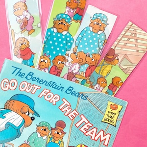 Könnte beinhalten: Ein Kinderbuch mit dem Titel "The Berenstain Bears Go Out for the Team" mit mehreren passenden Lesezeichen. Das Buchcover und die Lesezeichen zeigen Illustrationen von Bären beim Baseballspielen. Der Hintergrund ist ein helles Pink.
