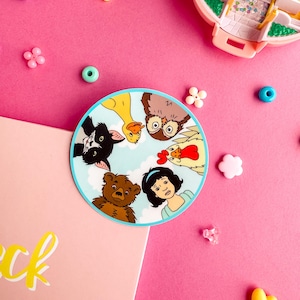Puede incluir: Una pegatina circular con una colorida ilustraci&oacute;n de un gato blanco y negro, un pato amarillo, un b&uacute;ho, un gallo, un oso pardo y una persona de pelo negro mirando hacia abajo desde un cielo azul claro lleno de nubes blancas.