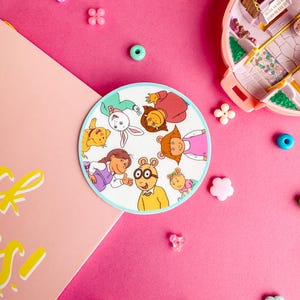 May include: A round sticker featuring the characters from the animated television series Arthur. The characters are arranged in a circle and include Arthur, Buster, Francine, Muffy, D.W., and Kate. The sticker is on a pink background with colorful beads and other decorative items.