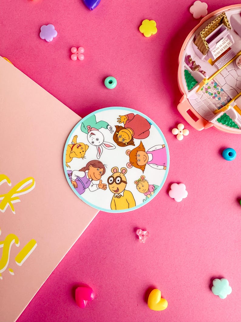 May include: A round sticker featuring the characters from the animated television series Arthur. The characters are arranged in a circle and include Arthur, Buster, Francine, Muffy, D.W., and Kate. The sticker is on a pink background with colorful beads and other decorative items.