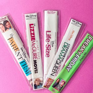 May include: Five Disney VHS tapes in clear plastic sleeves are arranged on a pink background. Titles include "The Parent Trap," "The Lizzie McGuire Movie," "Life-Size," "Confessions of a Teenage Drama Queen," and "Freaky Friday."