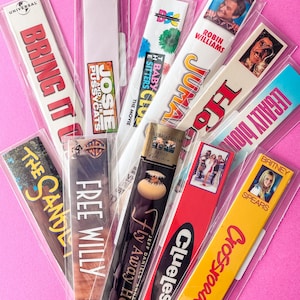 Puede incluir: Una colección de marcadores de cintas VHS vintage con títulos de películas como "Bring It On", "Josie and the Pussycats", "Jumanji" y "Legally Blonde". Cada marcador está en una funda de plástico transparente, con arte de películas.