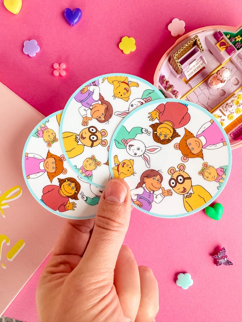 May include: Three round stickers featuring characters from the animated television series Arthur. The stickers are white with a blue border and feature the characters Arthur, D.W., Buster, Francine, Muffy, and the Brain.