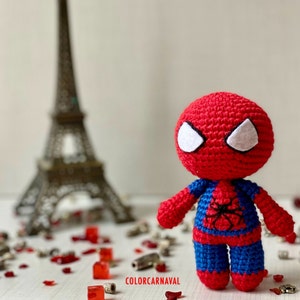 Puede incluir: Un juguete de Spiderman de crochet rojo y azul con ojos blancos. El juguete est&aacute; de pie sobre una superficie blanca con art&iacute;culos decorativos rojos y plateados.