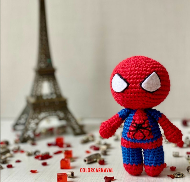 Puede incluir: Un juguete de Spiderman de crochet rojo y azul con ojos blancos. El juguete est&aacute; de pie sobre una superficie blanca con art&iacute;culos decorativos rojos y plateados.