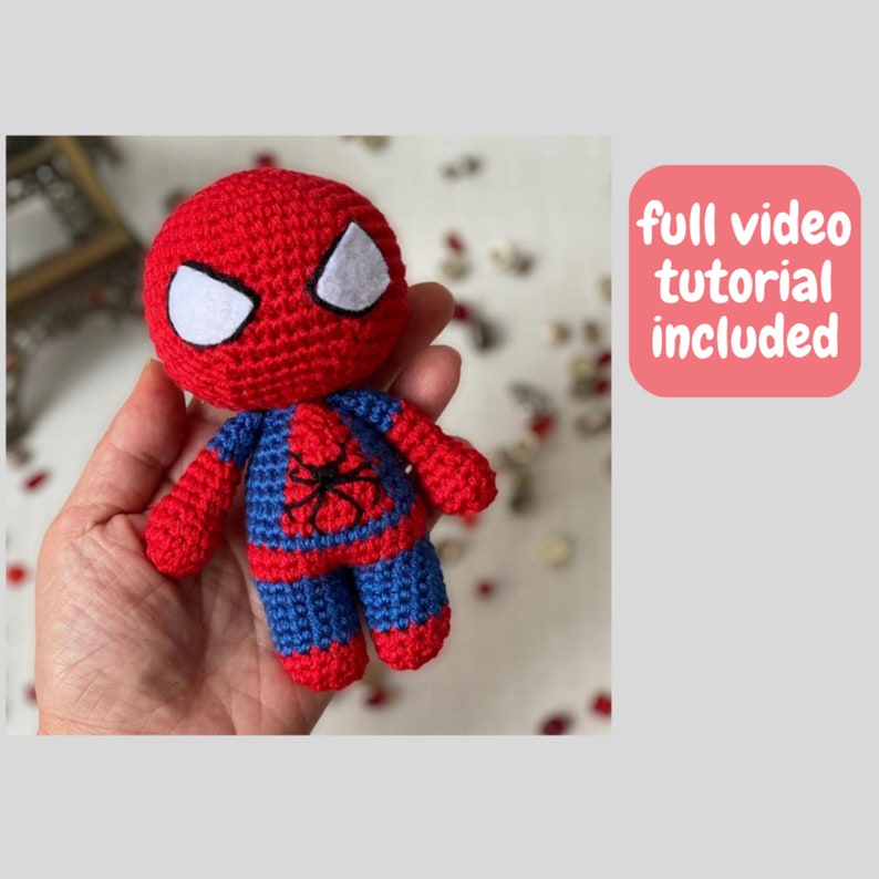Puede incluir: Un juguete de amigurumi Spiderman de crochet rojo y azul con ojos blancos. El juguete est&aacute; siendo sostenido en una mano. El texto "tutorial de video completo incluido" est&aacute; en un cuadro rosa.