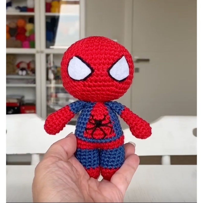 Puede incluir: Un juguete de Spiderman de crochet rojo y azul con ojos blancos. El juguete tiene los brazos extendidos y un dise&ntilde;o de ara&ntilde;a negra en el pecho.