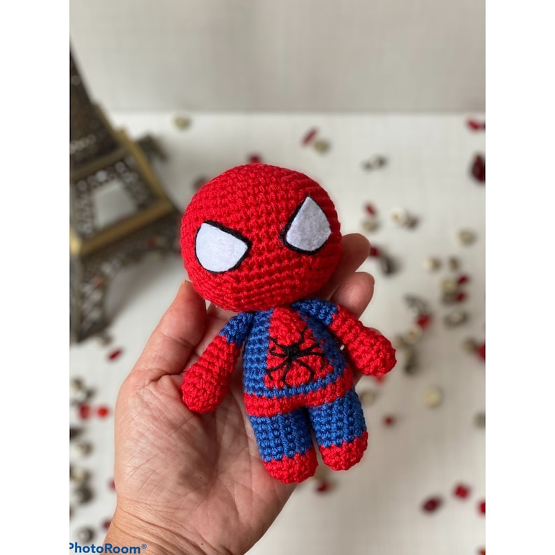 Puede incluir: Un juguete de Spiderman de crochet rojo y azul con ojos blancos. El juguete mide aproximadamente 15 cm de alto y tiene los brazos extendidos.