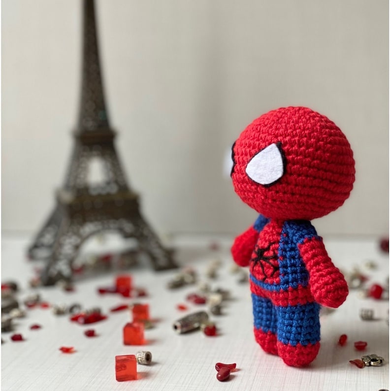 Puede incluir: Un juguete de Spiderman de crochet rojo y azul con ojos blancos. El juguete est&aacute; de pie sobre una superficie blanca con corazones rojos y blancos.
