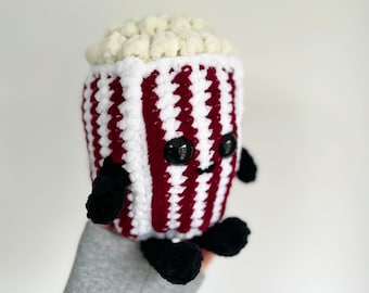 May include: A handmade, plush popcorn bucket with a smiling face, black eyes, and black arms and feet. The bucket is white with burgundy stripes and filled with fluffy, off-white popcorn. A cute, whimsical, and unique item.