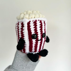 May include: A handmade, plush popcorn bucket with a smiling face, black eyes, and black arms and feet. The bucket is white with burgundy stripes and filled with fluffy, off-white popcorn. A cute, whimsical, and unique item.