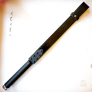 May include: A black leather riding crop with a long, rectangular handle and a rounded end. The handle is attached to a wide, black leather strap with a buckle. The strap has several holes and is secured with a metal rivet.