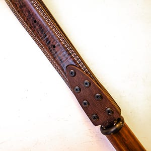 Pode incluir: Close-up de um cinto de couro castanho com costuras brancas e rebites de metal escuro. O cinto tem uma aparência texturizada e uma pega de madeira. O design é angulado, mostrando os detalhes do artesanato.