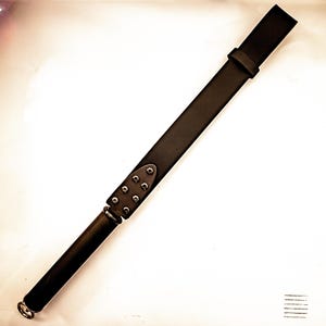 May include: A black leather riding crop with a rounded handle and a flat, rectangular end. The crop features a series of metal rivets and a metal ring at the base of the handle. The crop is set against a white background.