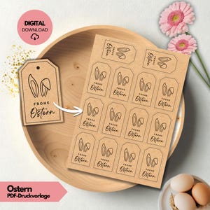Puede incluir: Etiquetas de regalo de Pascua de papel kraft con orejas de conejo y las palabras "Frohe Ostern" impresas en negro. Las etiquetas están dispuestas en una hoja. La imagen incluye flores rosas y huevos.