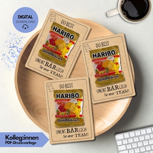 Pode incluir: Três cartões com embalagens de gomas Haribo são dispostos em uma bandeja de madeira. Cada cartão tem o texto "DU BIST HARIBO Goldbären UNENT BÄRLICH für unser TEAM!". Um ícone de download digital está no canto superior esquerdo.