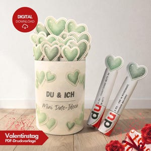 Könnte beinhalten: Ein zylindrischer Behälter mit grünen herzförmigen Aufsätzen, beschriftet mit "DU & ICH Mini Date-Ideen". Zwei weiße und rote Röhren mit Herzaufsätzen sind ebenfalls sichtbar. Das Bild enthält den Text "DIGITAL DOWNLOAD" und "Valentinstag PDF-Druckvorlage."