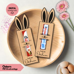 Könnte beinhalten: Oster-Themen-Digital-Download mit zwei hasenförmigen Haltern mit Schokoriegeln. Die Halter sind aus braunem Karton und haben schwarze Hasenohren. Der Text "Gruss vom Osterhasen" ist auf den Haltern aufgedruckt. Das Bild enthält auch rosa Blumen und Eier.
