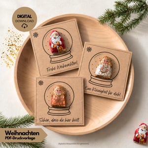 Könnte beinhalten: Drei quadratische Karten aus Kraftpapier mit schwarzen Strichzeichnungen von Schneekugeln, in denen jeweils eine Schokoladenfigur steckt. Die Karten tragen die deutschen Texte Frohe Weihnachten, Eine Kleinigkeit für dich und Schön, dass du hier bist.