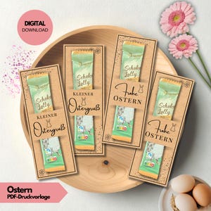 以下が含まれることがあります： 木製のトレイに、チョコレートバー付きのイースターカードがいくつかあります。「Frohe OSTERN」と「Kleiner Ostergruß」の文字とウサギのイラストが描かれています。ピンクの花と卵も画像に表示されています。