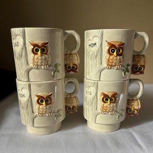 May include: Set of four ceramic mugs with owl designs. Each mug features a raised owl figure with brown, yellow, and white details, perched on a tree trunk. The mugs are labeled "DAD", "MOM", "SON", and "SIS" in heart-shaped designs.