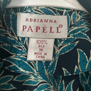 Puede incluir: Una camisa de seda azul y blanca con estampado de hojas tropicales. La camisa tiene una etiqueta que dice "Adrianna Papell" y "100% seda" con una etiqueta de talla 14. La camisa est&aacute; hecha en China.