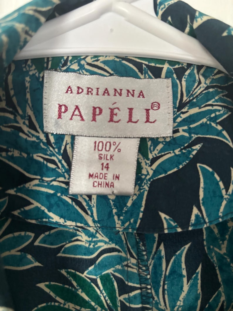 Puede incluir: Una camisa de seda azul y blanca con estampado de hojas tropicales. La camisa tiene una etiqueta que dice "Adrianna Papell" y "100% seda" con una etiqueta de talla 14. La camisa est&aacute; hecha en China.