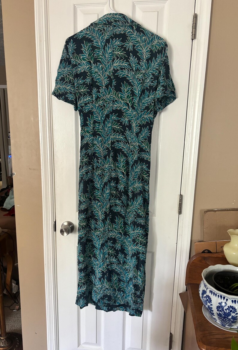Puede incluir: Un vestido largo de color azul oscuro con un estampado de hojas verdes y blancas. El vestido tiene mangas cortas y cuello.