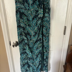 Puede incluir: Un vestido largo de color verde oscuro con un estampado de hojas blancas y verdes. El vestido tiene un lazo en la cintura y una abertura lateral.