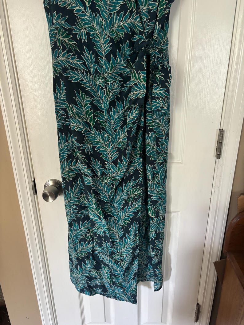 Puede incluir: Un vestido largo de color verde oscuro con un estampado de hojas blancas y verdes. El vestido tiene un lazo en la cintura y una abertura lateral.
