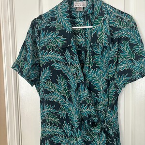 Puede incluir: Un vestido negro de envoltura con estampado de hojas verdes y blancas. El vestido tiene manga corta y cuello. La etiqueta de la marca dice "Adrienne Papell".