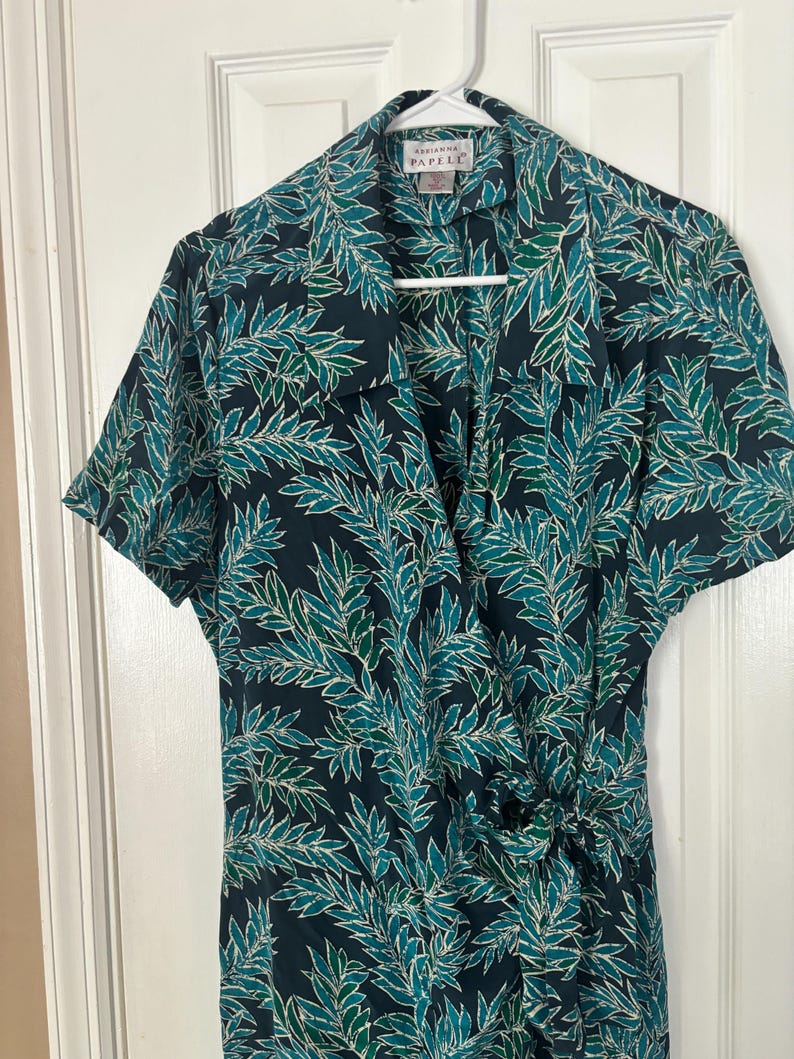 Puede incluir: Un vestido negro de envoltura con estampado de hojas verdes y blancas. El vestido tiene manga corta y cuello. La etiqueta de la marca dice "Adrienne Papell".