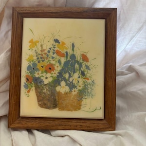 May include: A rectangular wall art piece featuring a ceramic tile with a floral illustration of yellow, orange, blue, and white flowers in two woven baskets, encased in a medium-toned wooden frame with a visible grain and a smooth finish.