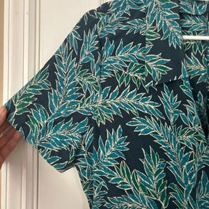 Puede incluir: Una camisa de manga corta azul oscuro con un estampado de hojas verdes y blancas. La camisa tiene cuello y cierre de botones en la parte delantera. La etiqueta de la camisa dice "Adriana Papell".