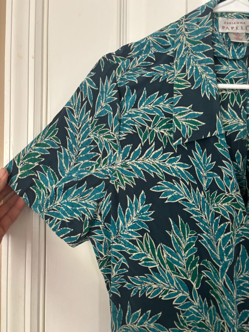 Puede incluir: Una camisa de manga corta azul oscuro con un estampado de hojas verdes y blancas. La camisa tiene cuello y cierre de botones en la parte delantera. La etiqueta de la camisa dice "Adriana Papell".